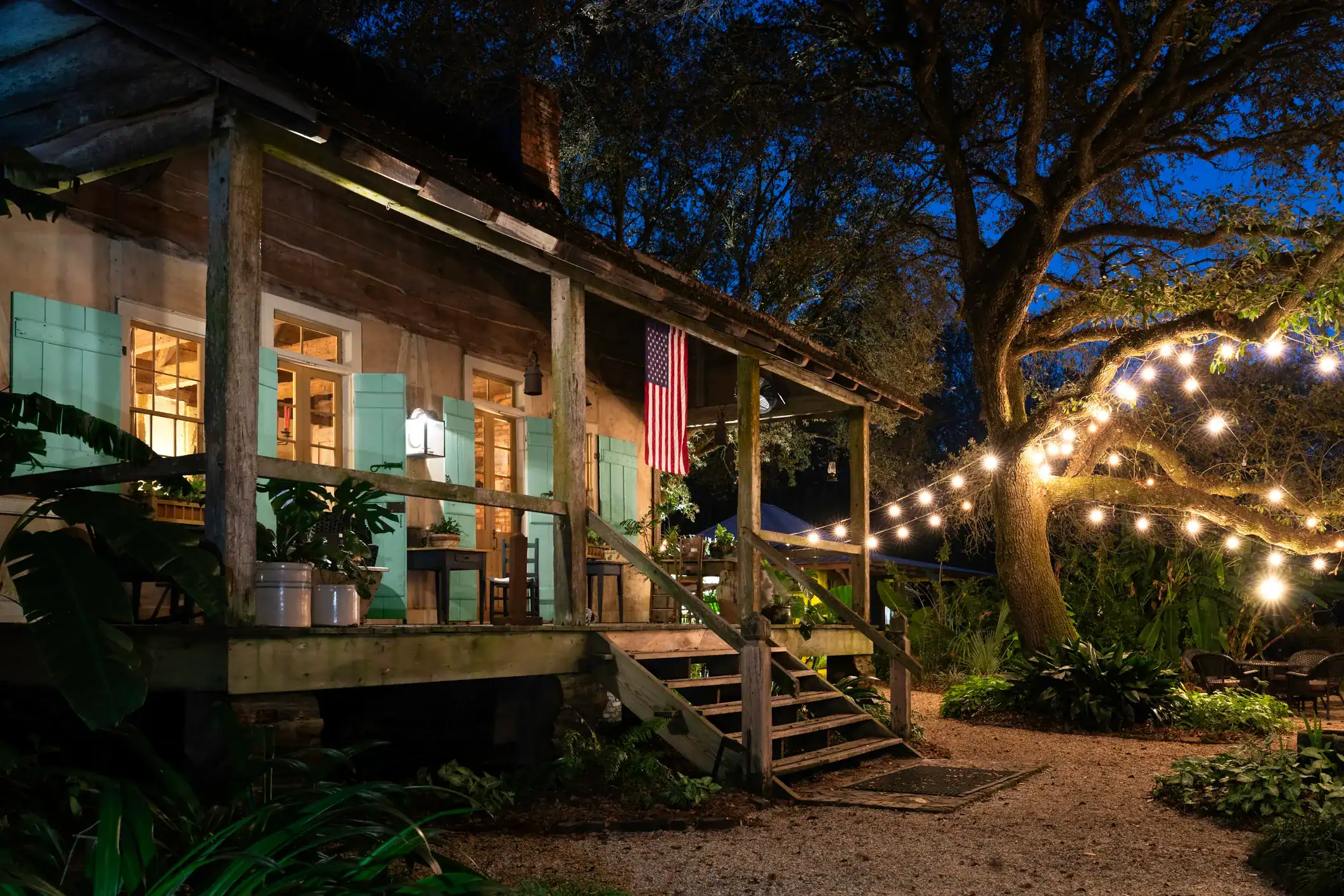 Cozy rustic cabin in Louisiana serving Cajun food illuminated by string lights at dusk, surrounded by lush greenery and tall trees.
