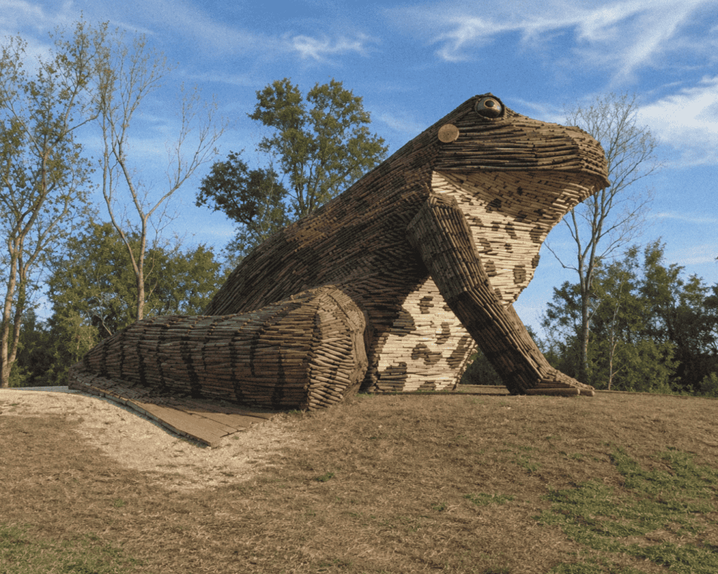 Large wooden frog bonfire sculpture outdoors on the levee with trees and blue sky in background.