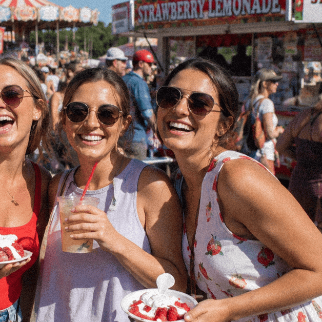 Three women enjoying strawberry lemonade at a Louisiana festival with food stalls in the background.