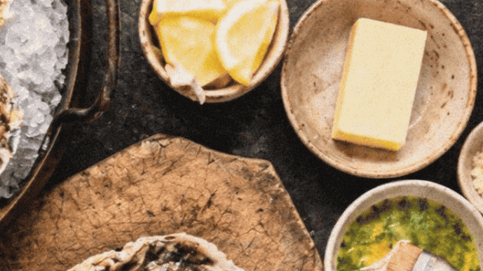 Oysters on a wooden board with lemon wedges, herbs, and sauces for making chargrilled oysters.